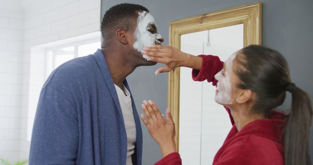 Image of happy diverse couple having fun applying cleansing face masks and laughing in bathroom. Happiness, love, self care, domestic life, and inclusivity concept.