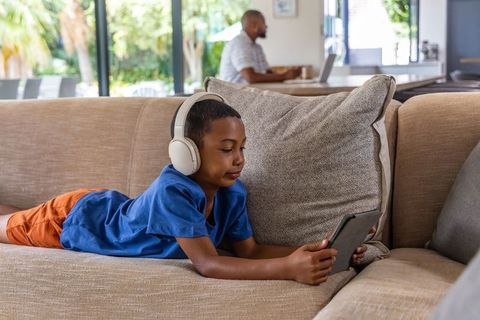 Boy lying on couch using tablet with headphones while father working from home