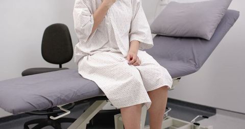 Female patient sitting on exam table wearing medical gown touching neck and resting on lap