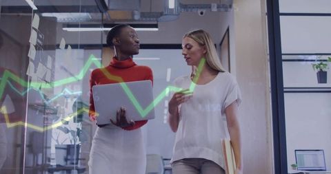 Walking businesswomen discussing data with laptop and glass charts in office hallway