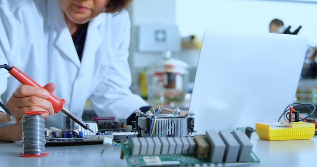 Caucasian woman works on electronic components in a lab. She's a technician focused on hardware repair, surrounded by tools and a laptop.