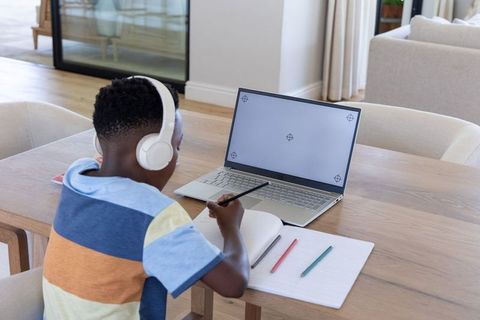 African american boy studying at home with laptop and headphones writing in notebook