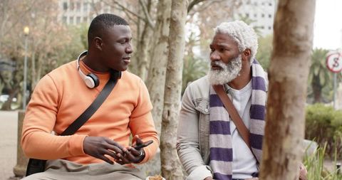 African american men having conversation on park bench while using smartphone