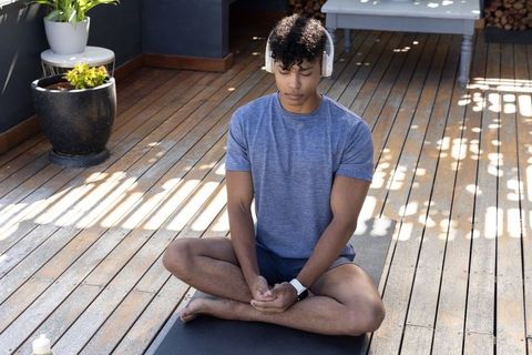 Young man meditating on sunlit terrace wearing headphones and smartwatch