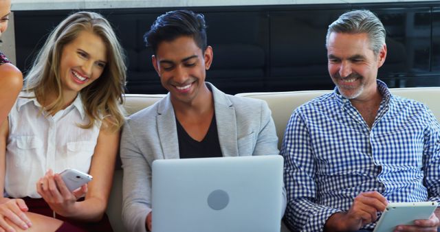 Group of professionals sitting on a couch, engaging with digital devices in a modern office environment. Suited for business, technology, and teamwork-related content such as articles, presentations, blogs, and social media posts to highlight collaboration and a multicultural workforce.