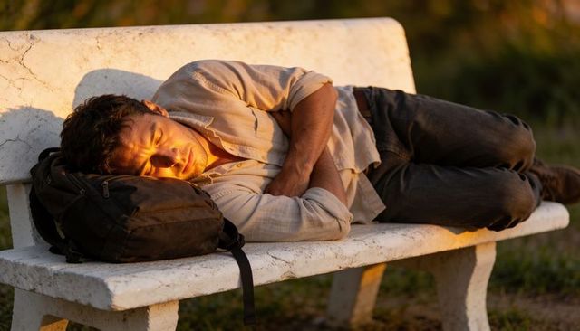 Sleeping man resting on white bench using backpack as pillow during golden hour