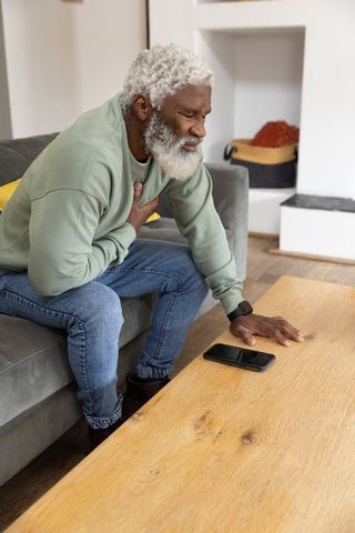Senior african american man clutching chest reaching for smartphone on table in distress