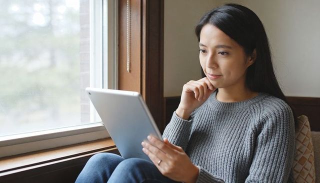 Woman relaxing on window seat reading tablet in cozy grey sweater, natural daylight