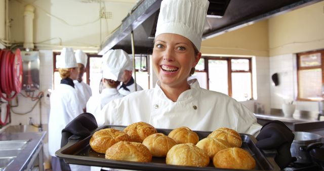 Female chef proudly presenting freshly baked bread rolls in a busy commercial kitchen environment. Ideal for use in articles about baking, culinary training, professional cooking, and kitchen team dynamics. Great visual for baking tutorials, restaurant marketing, and cookery content.