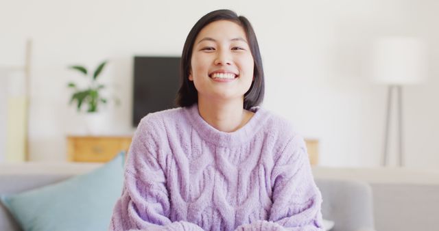 Young Asian woman sitting on a sofa in a cozy, inviting living room, wearing a purple sweater and smiling. Perfect for blogs and advertisements related to home comfort, cozy fashion, casual living, relaxation, and positive lifestyle. Ideal for use in social media marketing, lifestyle magazines, and home decor websites.