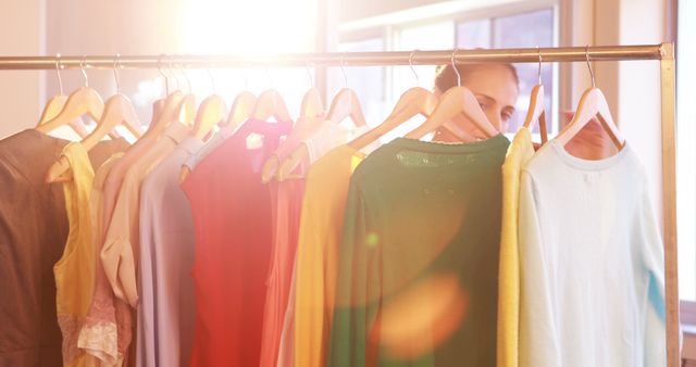 Individual browsing colorful clothes on rack in bright sunlit room, great for concepts like shopping, fashion, choices, stylish wardrobe, retail environments. Suitable for use in advertisements, blog posts, and social media promoting apparel and fashion.
