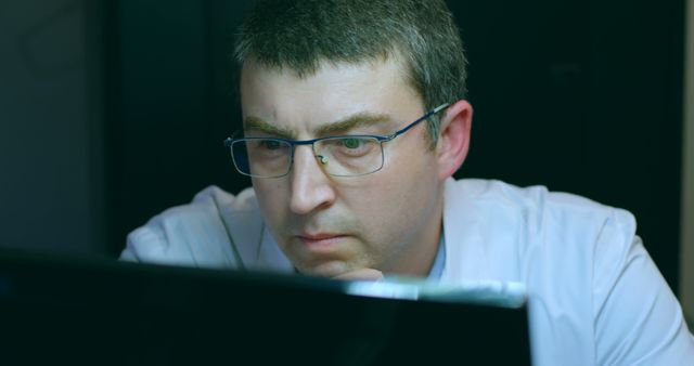 Middle-aged man wearing eyeglasses working on a laptop with focused expression. Suitable for illustrating concepts of hard work, dedication, late-night work, office environment, and technology use in professional settings.