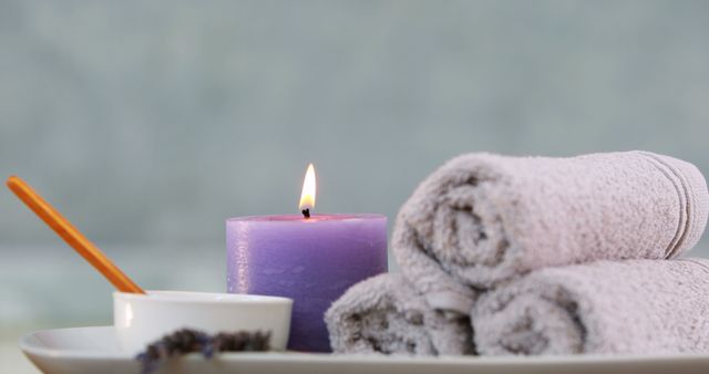 Purple candle burning next to three rolled towels and a small bowl on a tray. Ideal for use in promotions or blogs about spa services, wellness tips, or relaxation retreats.