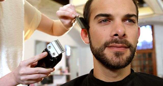 A man is receiving a haircut in a modern salon. The barber is using clippers and a comb to style his hair, creating a precise and clean look. This stock photo can be used for advertisements for barber shops, hair salons, and grooming products. Additionally, it is suitable for promoting services of professional hairstylists and personal care industries.