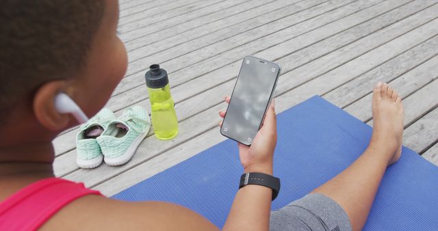 Image depicts a woman taking a break from her outdoor workout, sitting on a blue yoga mat, and using a smartphone. She is wearing athletic wear, listening with earbuds, and has a water bottle and sneakers nearby. Perfect for promoting fitness apps, healthy lifestyle blogs, or sports brands.