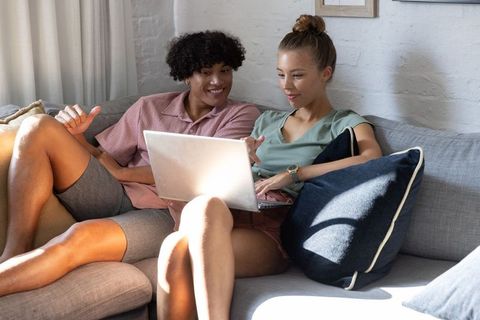 Couple relaxing on sectional sofa sharing laptop, browsing together in sunlit living room