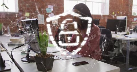 Woman working at desk holding coffee with digital cybersecurity shield overlay in office