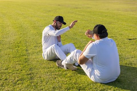 Two Teammates Sitting on Grass Signaling During Practice Wearing White Uniforms and Black Caps