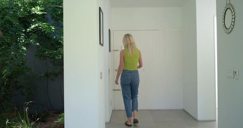 Blonde woman walking through minimalist foyer toward white paneled door with garden view