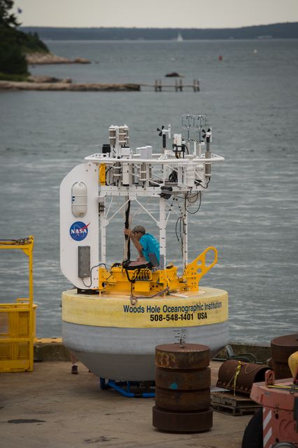 Senior engineer working on SPURS buoy on dock at Woods Hole, readying it for deployment. Research vessel Knorr standing by to transport buoy to Atlantic Ocean. Suitable for educational purposes, maritime research themed content, and showcasing preparations for scientific oceanographic studies.