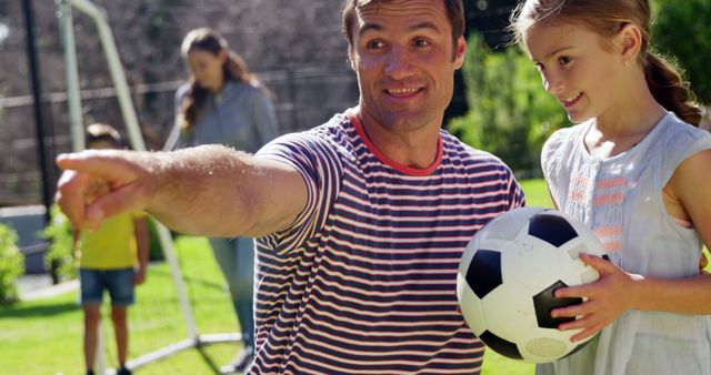 Father coaching daughter holding soccer ball outdoors. Great for depicting family bonding, sports training, and outdoor activities. Could be used in advertisements for sports equipment, family activities, and parenting content.