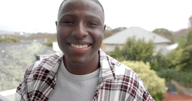 Young black man smiling warmly outdoors while wearing a casual plaid shirt and grey sweater. Ideal for themes of happiness, youth, confidence, casual fashion, and positive lifestyle.