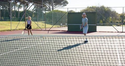 Asian man and woman practicing tennis on sunlit outdoor hard court with long shadows