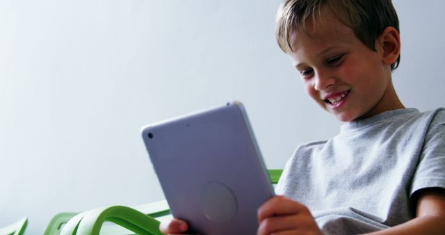 Young boy smiling and using a tablet while sitting in a green chair. Perfect for illustrating concepts related to digital learning, educational technology, childhood education, and modern classrooms.