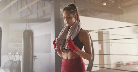 Female boxer pausing with towel beside ring and heavy bag showing focus and strength