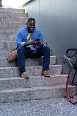 African american commuter sitting on stairs checking smartphone and smiling with bicycle