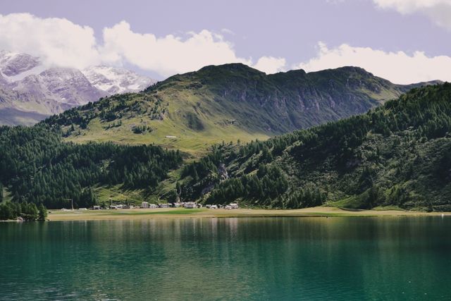 Snow-capped mountains in background overlook a tranquil lake bordered by lush green hills. Tiny rural village at the water's edge. Ideal for nature, travel, outdoors, and serenity themes, emphasizing scenic Alpine beauty and peaceful countryside retreats.