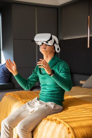 Asian man wearing vr headset on bed gesturing in minimalist room with mustard throw
