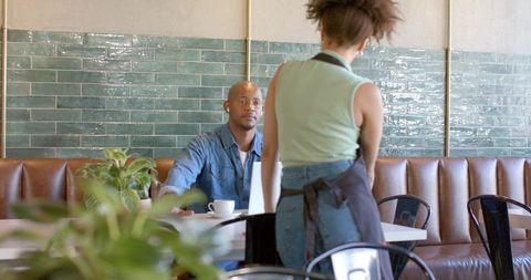 Server taking order from seated customer at laptop with coffee mug in sunlit café