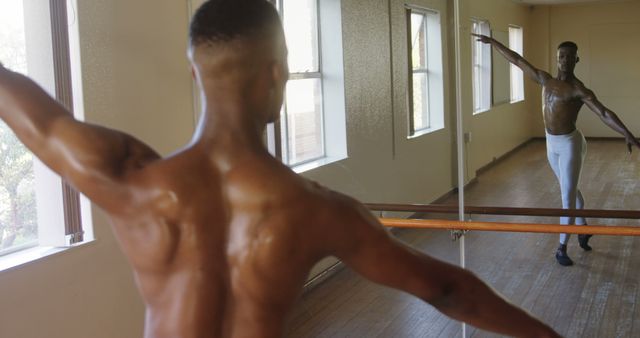Bare-chested male ballet dancer practicing in a sunlit studio standing before a mirrored wall, with arms outstretched. Suitable for themes related to dance, fitness, dedication, artistic expression, and ballet training.