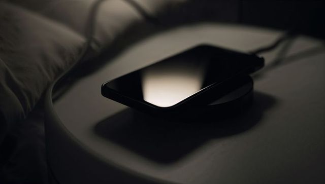 Black smartphone resting on wireless pad on bedside table at night, moody reflection
