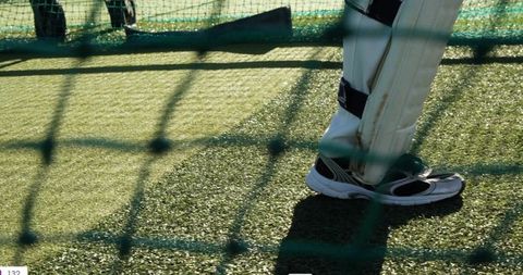 Cricket batter standing in leg pads and running shoes on artificial turf with net shadows