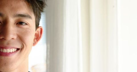 Smiling young asian man leaning by window with sheer curtains and natural light