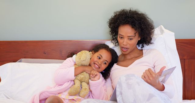 This image showcases a tender moment between a mother and daughter at bedtime. The mother reads a story while the daughter cuddles a teddy bear, highlighting family bonding and the comforting routines of childhood. This could be used for topics related to parenting tips, bedtime routines, family activities, and connections between parents and children.
