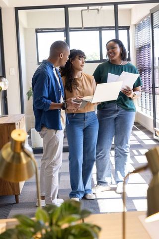 Diverse coworkers collaborating in sunlit modern office corridor with laptop and documents