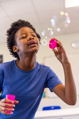 African american boy blowing bubbles in classroom playing with pink wand and bubble jar