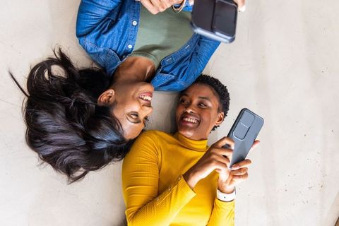 Two diverse women lying overhead smiling using smartphones taking selfie sharing moment