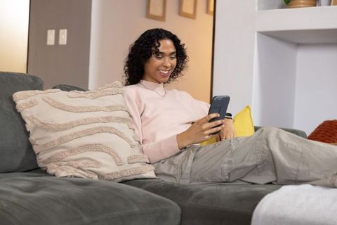 Non-binary adult relaxing on sofa scrolling smartphone in cozy modern living room