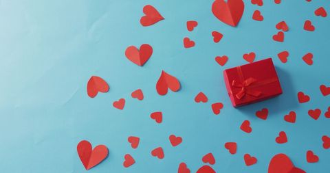 Red gift adorned with ribbon among scattered paper hearts on vibrant blue field. Evoking Valentine's Day ambiance and theme of romance and celebration. Ideal for advertisements, greeting cards, holiday flyers, and social media content promoting love-centric themes.