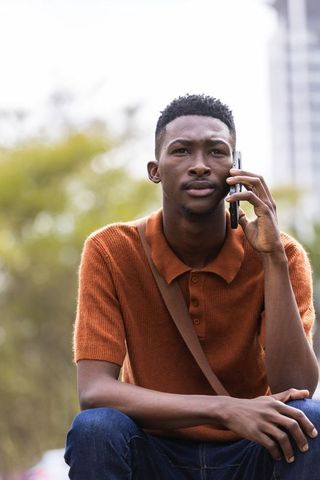 Thoughtful african man sitting in urban park talking on smartphone in rust polo portrait