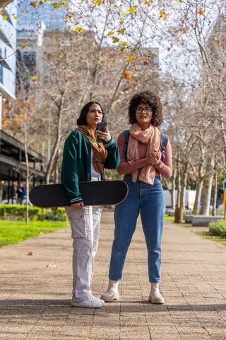 Diverse women standing on urban walkway holding skateboard and smartphone wearing scarves