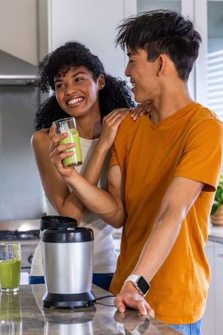 Diverse couple smiling and preparing green smoothies in bright modern kitchen
