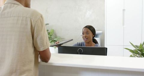 African american receptionist exchanging laptop with visitor at modern reception desk
