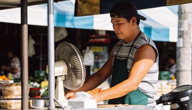 Market vendor arranging baked goods under striped canopy at sunlit street food stall