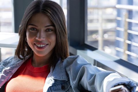 Smiling woman in denim jacket holding smartphone in sunlit modern lounge by windows
