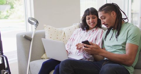 Biracial Couple on Couch Enjoy using phone and laptop Together, Crutch Nearby